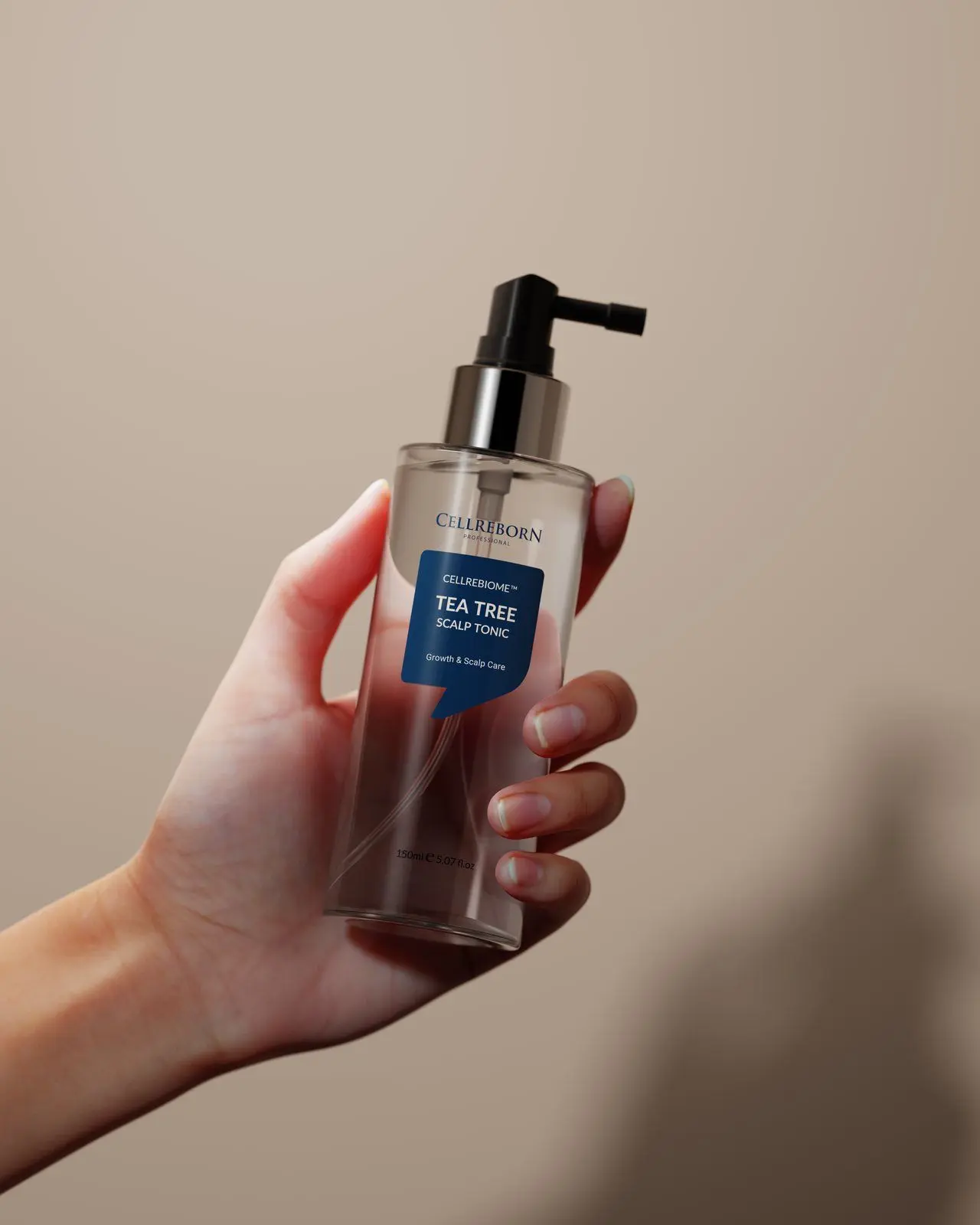 A hand holding a Cellrebiome Tea Tree Scalp Tonic bottle against a taupe backdrop